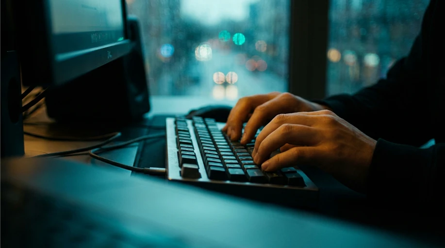 Mãos digitando em teclado à noite, iluminadas pela luz de uma tela